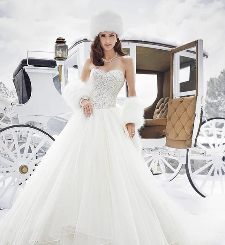 Bride in elegant white gown posing beside decorative white horse carriage in snowy setting